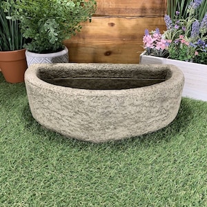 May include: A half-round concrete planter with a rough, textured surface. The planter is empty and sits on a green artificial grass surface.