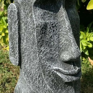 May include: A grey stone sculpture of a human head with a large nose and a closed mouth. The sculpture has a rough, textured surface and is sitting on a bed of green grass.