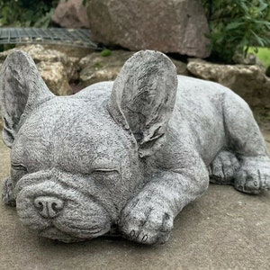 May include: A grey concrete sculpture of a French bulldog sleeping. The sculpture is detailed with realistic features, including wrinkles and folds in the dog's skin.