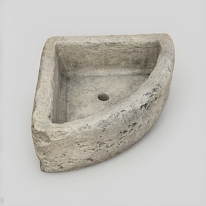 May include: A corner-shaped stone basin with a rough, textured exterior and a smooth interior surface. The basin features a single circular drainage hole centred at the bottom, with thick, weathered walls and a light grey, mottled stone finish.