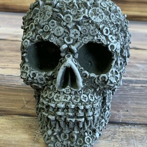 May include: A grey skull sculpture made of concrete and covered in screws and nuts.