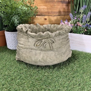 May include: A grey concrete planter shaped like a burlap sack with a rope tie. The planter is sitting on a green artificial grass surface.