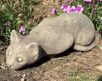 May include: A gray concrete cat statue lying on its side with its head turned to the left. The statue has realistic features, including eyes, a nose, and whiskers.