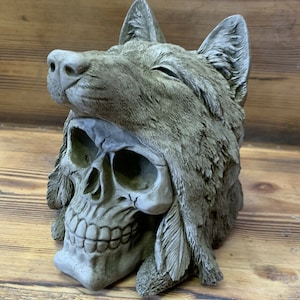 May include: A grey concrete sculpture of a wolf's head with a skull underneath. The wolf's head is tilted back and the skull is facing forward. The sculpture is detailed with feathers and fur.