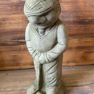 May include: A grey stone statue of a golfer wearing a hat and holding a golf club. The golfer is standing on a base with a golf ball.