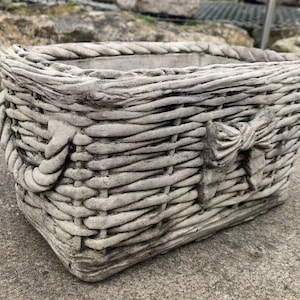May include: A grey concrete planter shaped like a woven basket with a bow detail. The planter is ideal for adding a rustic touch to any garden or home.
