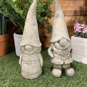 May include: Two grey concrete garden gnomes. The gnome on the left is wearing a dotted hat and a dress. The gnome on the right is wearing a hat and a jacket. Both gnomes are smiling.