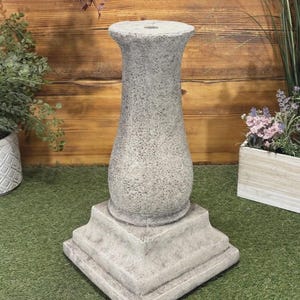 May include: A grey, textured pedestal with a square base and a fluted column. The pedestal is made of a stone-like material, suitable for displaying plants or decorative items. The background features wooden planks and greenery.