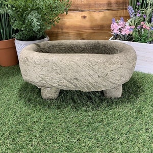 May include: A grey stone oval planter with a rough texture and three short legs. The planter is sitting on a green artificial grass surface.