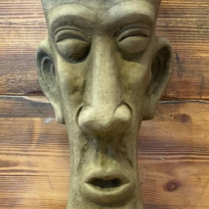May include: A grey concrete sculpture of a face with closed eyes and a large nose. The sculpture is a planter with a wide opening at the top.