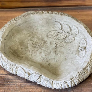 May include: A grey concrete bird bath with a leaf design around the rim and a leaf design in the centre.
