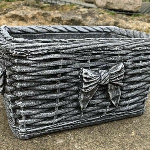May include: A grey concrete planter shaped like a woven basket with a large bow on the front.