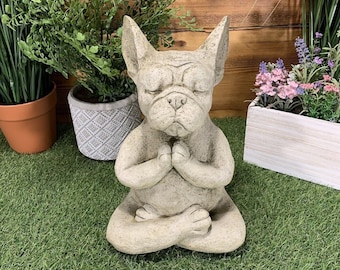 May include: A gray concrete statue of a French bulldog sitting in a yoga pose with its eyes closed. The statue is on a green artificial grass surface.