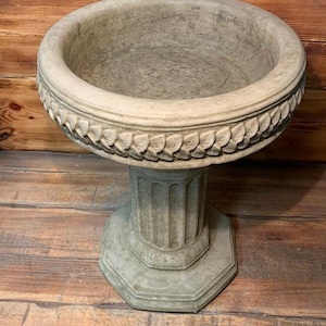 May include: A grey concrete bird bath with a decorative leaf design around the rim. The bird bath is on a pedestal with a fluted column.