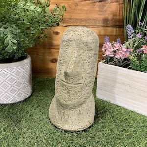May include: A grey stone sculpture of a face with a carved smile. The sculpture is sitting on a green artificial grass surface.