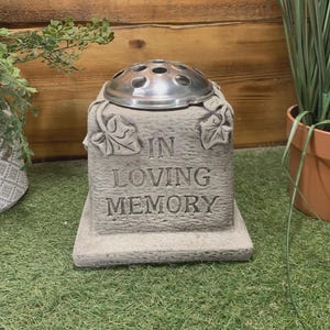 May include: A grey stone memorial flower vase with a silver metal flower holder. The vase is inscribed with the words "IN LOVING MEMORY". The vase has leaf designs and sits on a rectangular base.