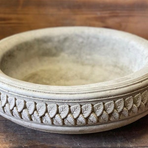May include: A round, grey concrete planter with a decorative leaf design around the rim.