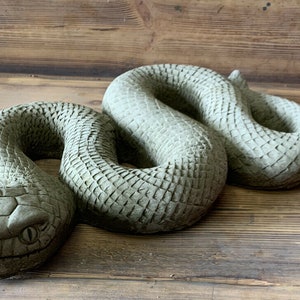 May include: A grey concrete snake sculpture with a detailed textured surface, coiled in a realistic pose. The snake has a defined head with eyes and scales.