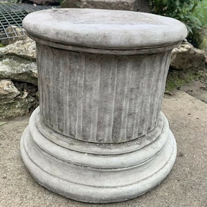 May include: A grey concrete pedestal with a fluted column and a rounded top. The pedestal is ideal for displaying plants, sculptures, or other decorative items.