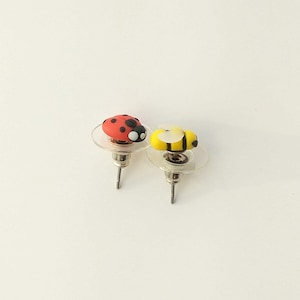 May include: A pair of stud earrings featuring a red ladybug and a yellow bee. The earrings are made of polymer clay and have silver posts.