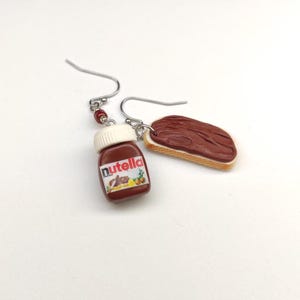 Pode incluir: Um par de brincos pendentes com um pote de Nutella em miniatura e uma fatia de pão com pasta de chocolate. Os brincos são feitos de argila polimérica e têm ganchos prateados.