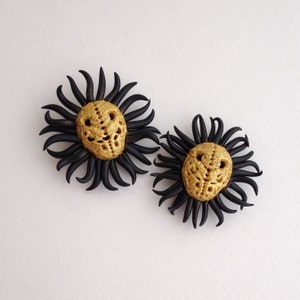 May include: Two handmade, artistic earrings. Each earring features a gold-colored, textured skull design at the center, surrounded by black, curved, petal-like elements. The earrings are set against a white background.