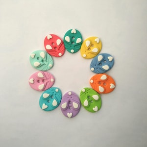 May include: A collection of colorful, oval-shaped buttons arranged in a circle. Each button features a different color, including red, green, yellow, and blue, with white decorative accents. The buttons are likely for crafts or sewing projects.