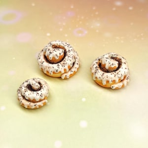 Könnte beinhalten: Drei Miniatur-Zimtschnecken mit weißem Zuckerguss und dunklen Streuseln. Die Brötchen sind auf einem hellgelben und grünen Farbverlauf angeordnet. Der Zuckerguss ist über die Brötchen geträufelt, und die Streusel sorgen für Textur.