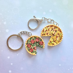 Peut inclure: Deux porte-clés miniatures en forme de part de pizza. L'un présente une pizza avec des tranches de tomates rouges et des herbes vertes. L'autre a une pizza avec du fromage jaune et des garnitures rouges. Les deux ont une chaîne et un fermoir argentés.