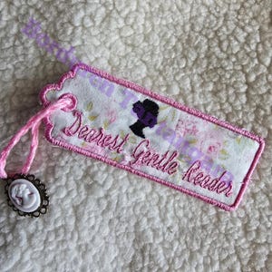 Puede incluir: Marcapáginas bordado con borde rosa y estampado floral. El marcapáginas presenta el texto "Dearest Gentle Reader" y una silueta de mujer. Se adjuntan un cordón rosa y un dije de camafeo.