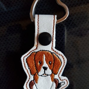 Op de afbeelding: Wit leren sleutelhanger met een bruin en wit geborduurd beagle-hond ontwerp. De sleutelhanger heeft een hartvormige metalen ring.