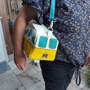 Op de afbeelding: Een witte en gele schoudertas met een blauw en wit busontwerp. De tas heeft een turquoise band en een witte gesp. De tas heeft de tekst "LA 242" aan de voorkant.