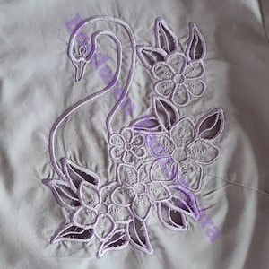 Può includere: Un ricamo di colore lilla chiaro che rappresenta un cigno con dei fiori su un tessuto grigio chiaro.