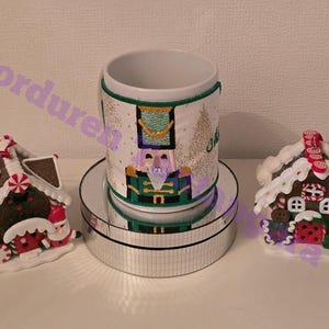 May include: A white ceramic mug with a nutcracker design, flanked by two gingerbread house ornaments. The mug features a green and gold trim and a festive illustration. The gingerbread houses have white icing and candy decorations.