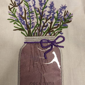 Op de afbeelding: Borduurwerk van een paarse lavendelboeket in een glazen pot met een paarse strik om de deksel. De pot heeft een bruin label met de tekst "CREA LINDA" erop.