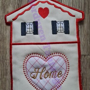 May include: A white fabric house-shaped wall hanging with red trim. The house has two windows and a red heart on the front. The bottom of the house has a red heart with the word "Home" embroidered in gold.