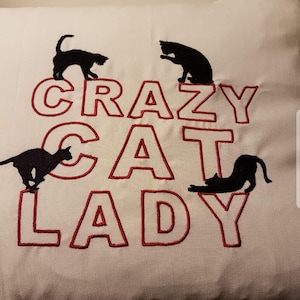 Op de afbeelding: Wit kussen met de geborduurde tekst "CRAZY CAT LADY" in rood, met zwarte kat silhouetten. De katten staan in verschillende poses, wat een speelse touch aan het ontwerp geeft. Een decoratief item voor kattenliefhebbers.
