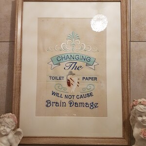 Op de afbeelding: Ingelijst kunstwerk met een lichtbruine houten lijst. Het kunstwerk toont geborduurde tekst met de tekst "CHANGING The TOILET PAPER WILL NOT CAUSE Brain Damage" in blauw en wit garen. De achtergrond is een beige stof.