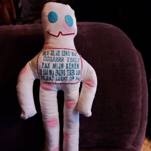 Op de afbeelding: Een witte stoffen pop met roze stippen en blauwe ogen. De pop heeft een rood gestikte glimlach en een blauwe geborduurde boodschap op zijn borst die luidt: "ALS JE JE EENS WAT GGGRRRR VORLT EAK MIJN BENEN EN SLAMDA ENGERS TEGEN AAN 20.DAT LUCHT OP HO"
