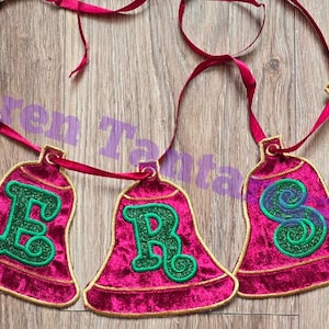 Op de afbeelding: Een feestelijke banner met vijf klokvormige ornamenten. Elke bel is diep magenta van kleur met een gouden rand en een groene letter. De bellen zijn verbonden door een magenta lint.