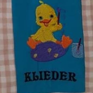 Op de afbeelding: Geborduurde turquoise stof met een gele eend op een paars ei. Het eendje heeft oranje voetjes en een rode snavel. Het woord "KLIEDER" is in het zwart geborduurd aan de onderkant. De achtergrond is een roze en wit geruit patroon.