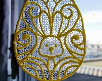 Op de afbeelding: Een geel en wit gehaakt paasei-ornament met een konijnenontwerp.