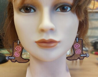 May include: A pair of brown cowboy boot earrings with pink flower accents. The earrings are hanging from a mannequin's ears.