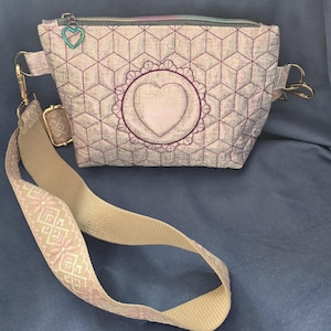 May include: A crossbody bag featuring a light-colored fabric with a dark geometric cube pattern and a central embroidered heart within a scalloped circular frame. It includes a rainbow-colored zipper, a heart-shaped charm, and a wide beige strap.