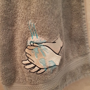 Könnte beinhalten: Graues Handtuch mit einem gestickten Design von zwei Händen, die sich mit Wasser aus einem Wasserhahn waschen.
