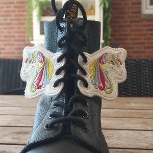 May include: Black leather boot with embroidered unicorn wing patches. The patches are white with a colorful rainbow mane and tail. The boot has black laces and is sitting on a wooden surface.