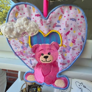 Op de afbeelding: Een hartvormige, roze en blauwe geborduurde decoratie met een roze teddybeer, een witte wolk en een snoeppatroon. De decoratie heeft een blauwe rand en een gele accent, hangend aan een roze lint.