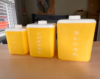 May include: Three yellow ceramic canisters with white lids and white lettering. The canisters are labeled "Tea", "Coffee", and "Sugar".