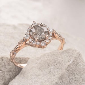 Può includere: Un anello di fidanzamento in oro rosa con un diamante grigio a forma di pera circondato da un alone di diamanti più piccoli. La fascia dell'anello è decorata con delicati dettagli a forma di foglie.