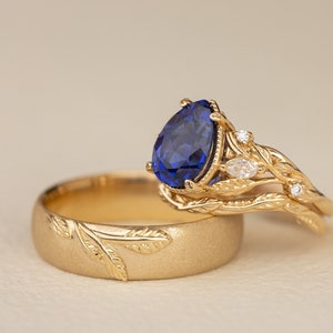 May include: A pair of gold wedding bands with a leaf design. The engagement ring features a pear-shaped blue sapphire and two small diamonds.